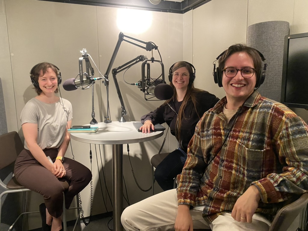Madeleine in a podcast booth with two of her interns, all three smiling at the camera