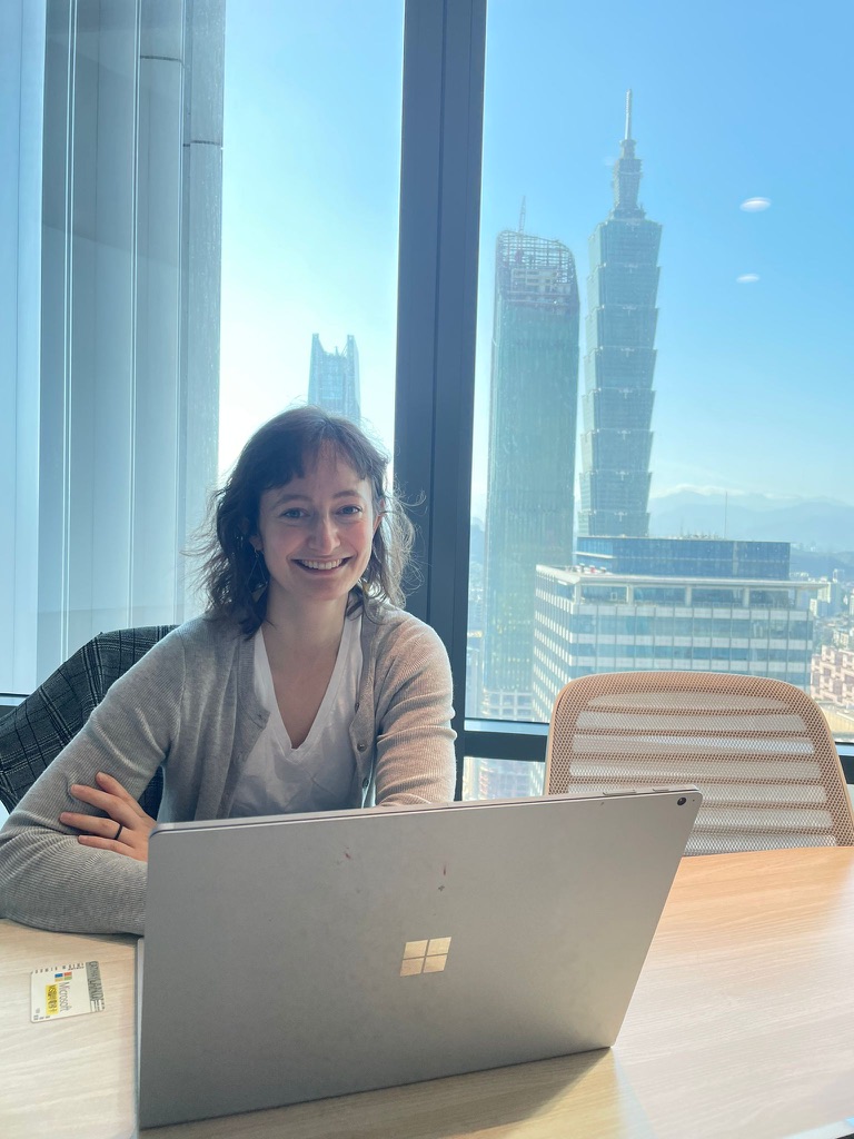 Madeleine smiling at her laptop with Taipei 101 visible through the window